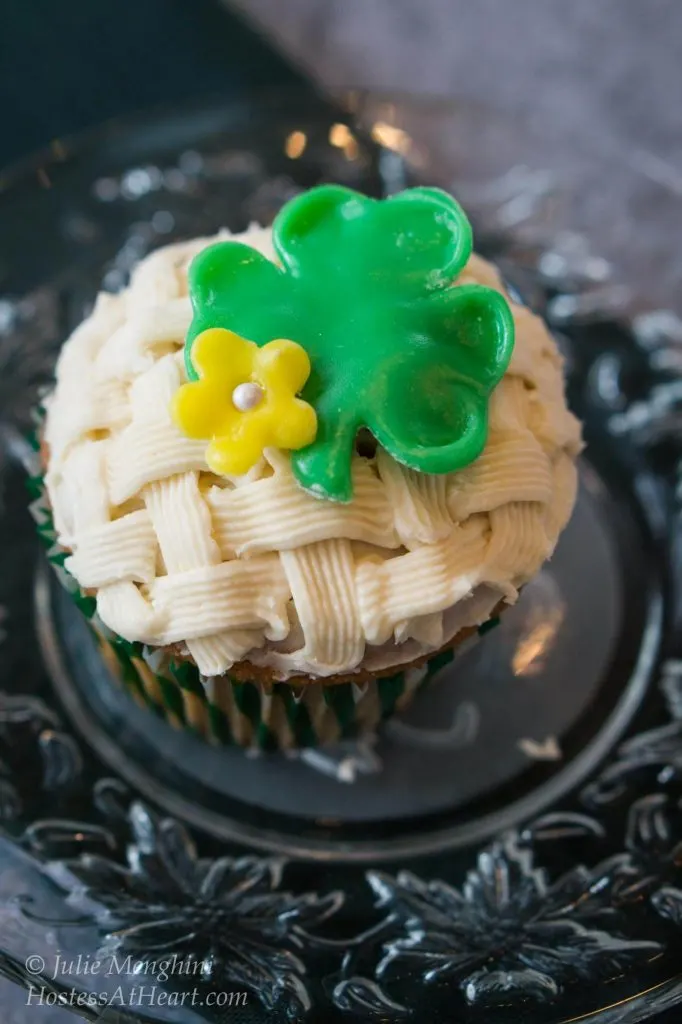 Cupcake decorated with a basketweave pattern. A green Shamrock and flower sit on the top. The cupcake sits on a metal plate.