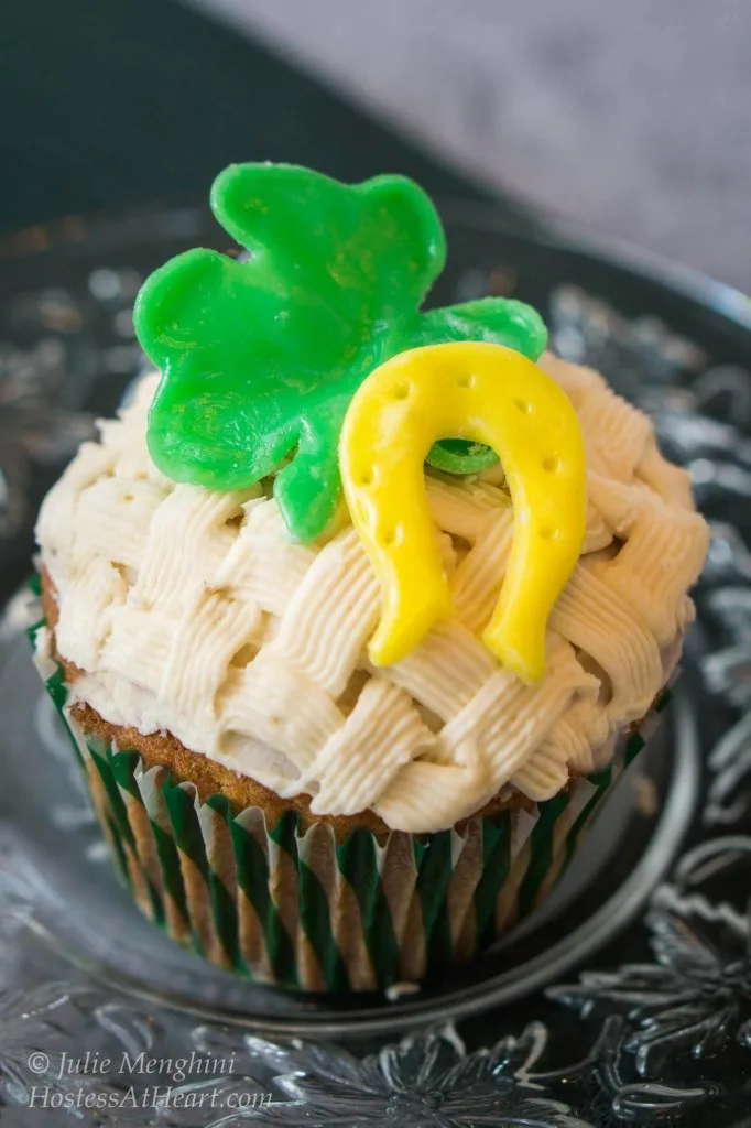 Cupcake decorated with a basketweave pattern. A green Shamrock, horseshoe, and flower sit on the top. The cupcake sits on a metal plate.