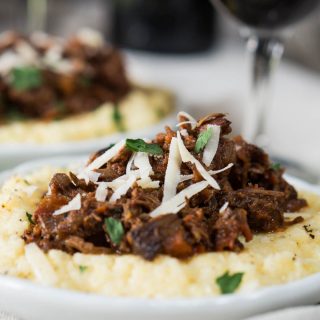 Side view of a lamb Ragu on a pile of cheesy polenta and garnished with shredded cheese and chopped parsley sitting on a white plate. A second plate and a wine glass sit in the background.