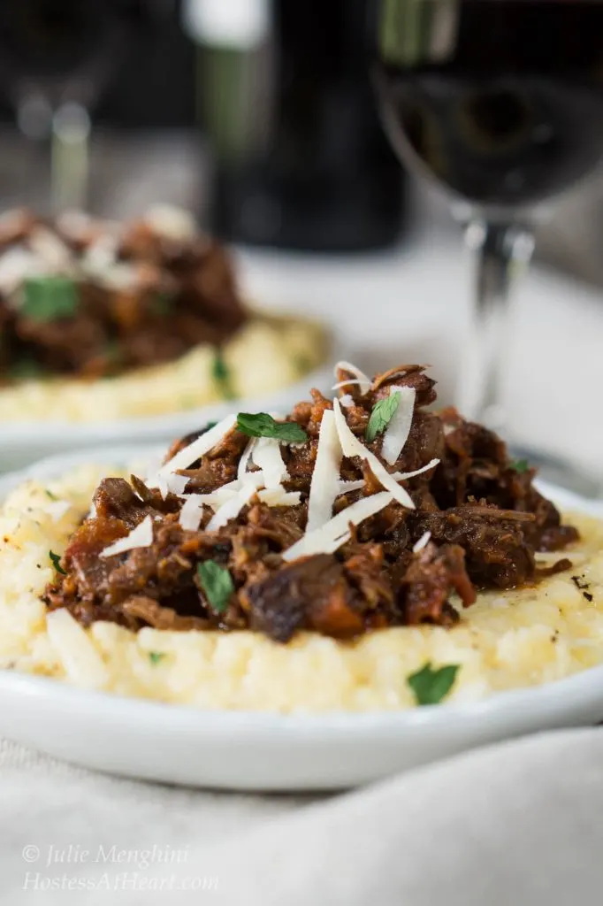 Side view of a lamb Ragu on a pile of cheesy polenta and garnished with shredded cheese and chopped parsley sitting on a white plate. A second plate and a wine glass sit in the background.
