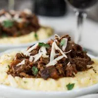 Side view of a white plate filled with cheesy polenta and topped with a mound of lamb Ragu garnished with parmesan cheese. A second plate sits in the background next to a glass of wine.