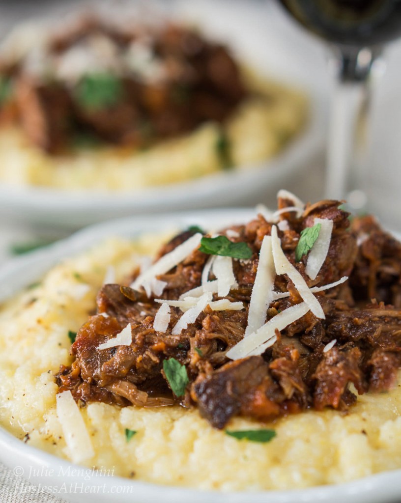 Side view of a white plate filled with cheesy polenta and topped with a mound of lamb Ragu garnished with parmesan cheese. A second plate sits in the background next to a glass of wine.