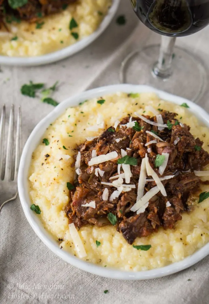 Top-down view of a white plate filled with cheesy polenta and topped with a mound of lamb Ragu garnished with parmesan cheese. A second plate sits in the background next to a glass of wine.