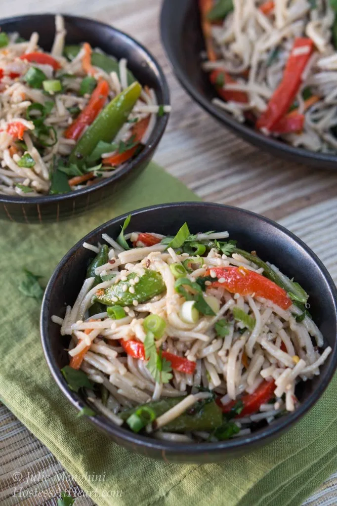 Top-down view of two bowls of black bowls filled with soba noodles with Asian flavors stuffed with red peppers and garnished with sesame seeds and cilantro. A larger serving bowl sits in the background.