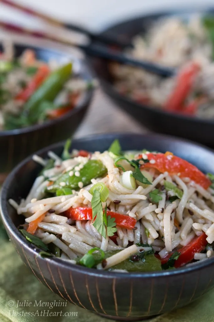 A black bowl filled with soba noodles with Asian flavors stuffed with red peppers and garnished with sesame seeds and cilantro. A second bowl sits in the background next to a serving bowl.