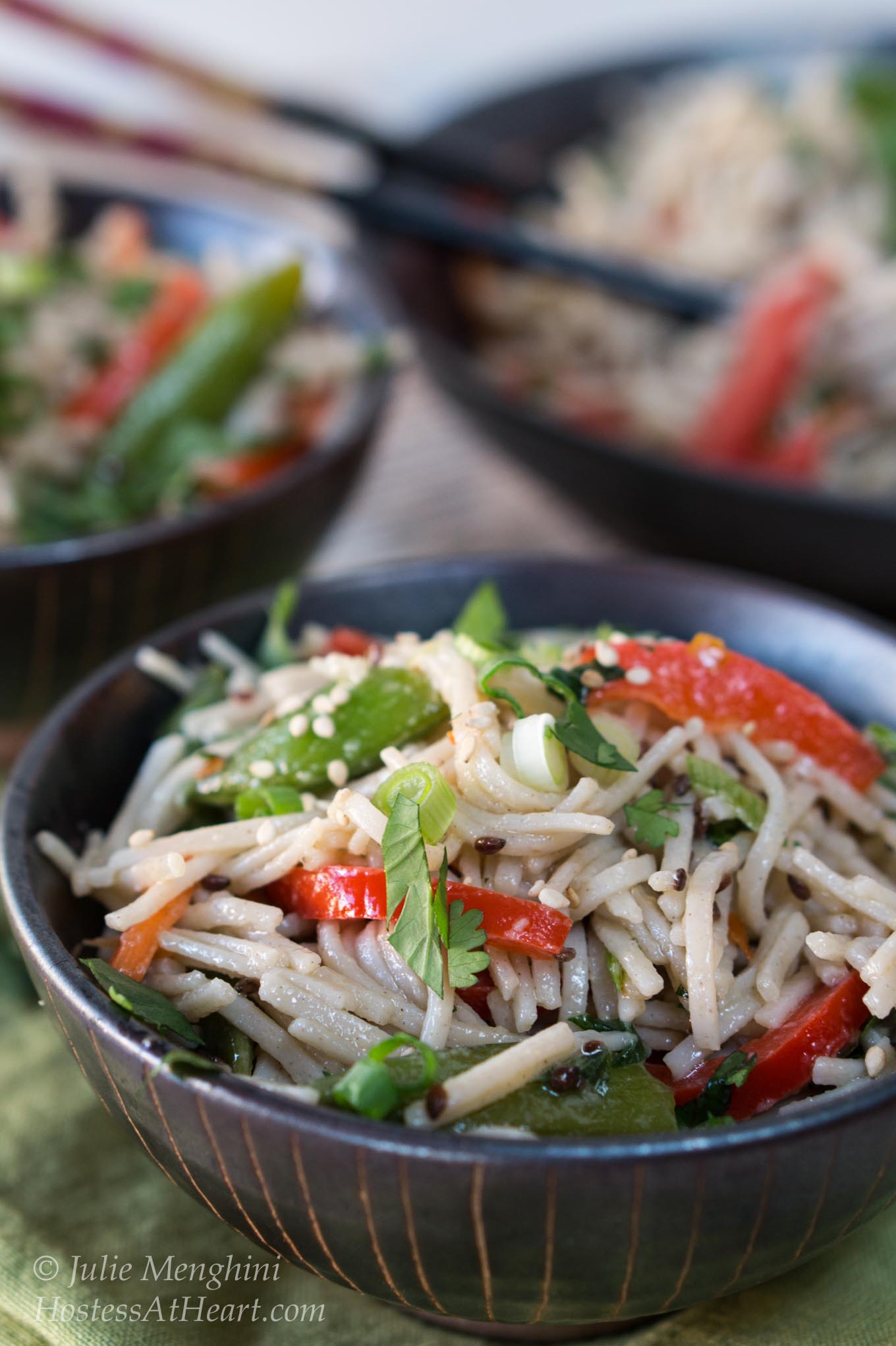 A black bowl filled with soba noodles with Asian flavors stuffed with red peppers and garnished with sesame seeds and cilantro. A second bowl sits in the background next to a serving bowl.