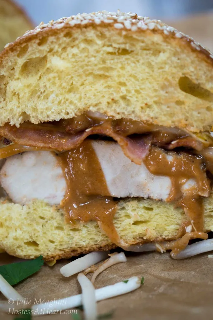 Close up table view of half a chicken sandwich with sliced bacon and with peanut sauce running out of it. Bean sprouts are scattered in the forefront.