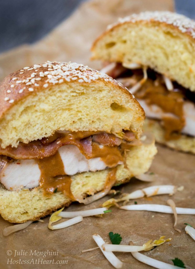 Side view of a chicken sandwich on sesame seed brioche bun that\'s been cut in half. Peanut sauce is drizzling out of the sandwich.