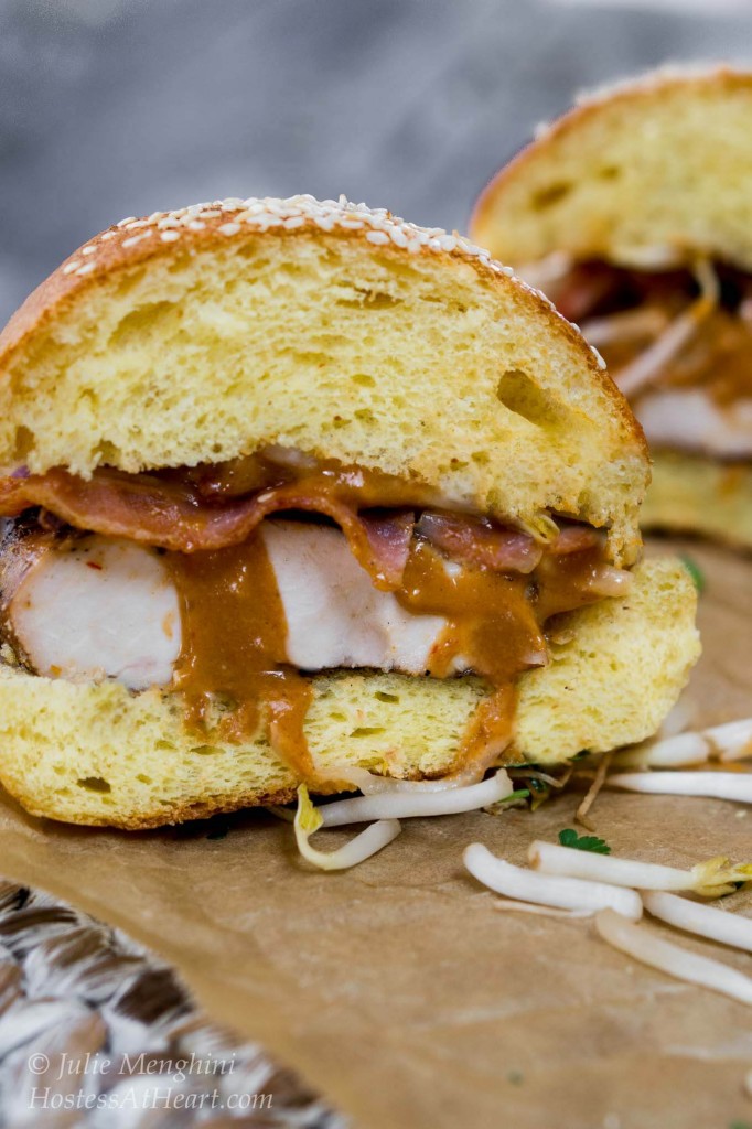 Table view of a half of chicken sandwich with peanut sauce and bacon on a sesame brioche bun. Bean sprouts are scattered around the sandwich.