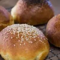 Angled view of brioche buns over a cooling rack and topped with sesame seeds or poppy seeds. Some are left plain.