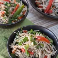 Top-down view of two bowls of black bowls filled with soba noodles with Asian flavors stuffed with red peppers and garnished with sesame seeds and cilantro. A larger serving bowl sits in the background.