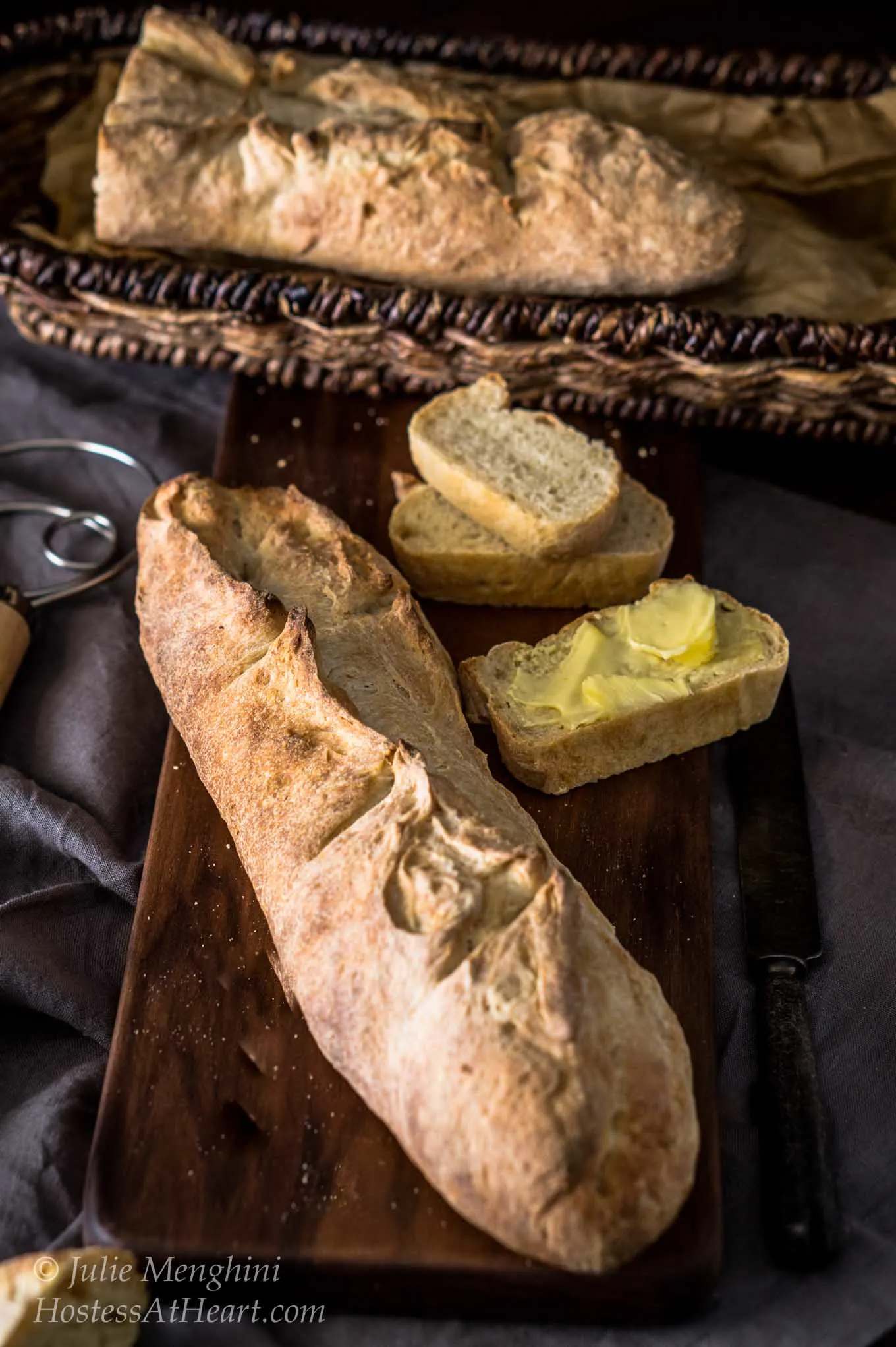 Top-down view of a loaf of a Garlic Batard sitting on a wooding cutting board next to cut slices slathered with butter. A blue napkin sits to the side and a basket of bread sits in the background.