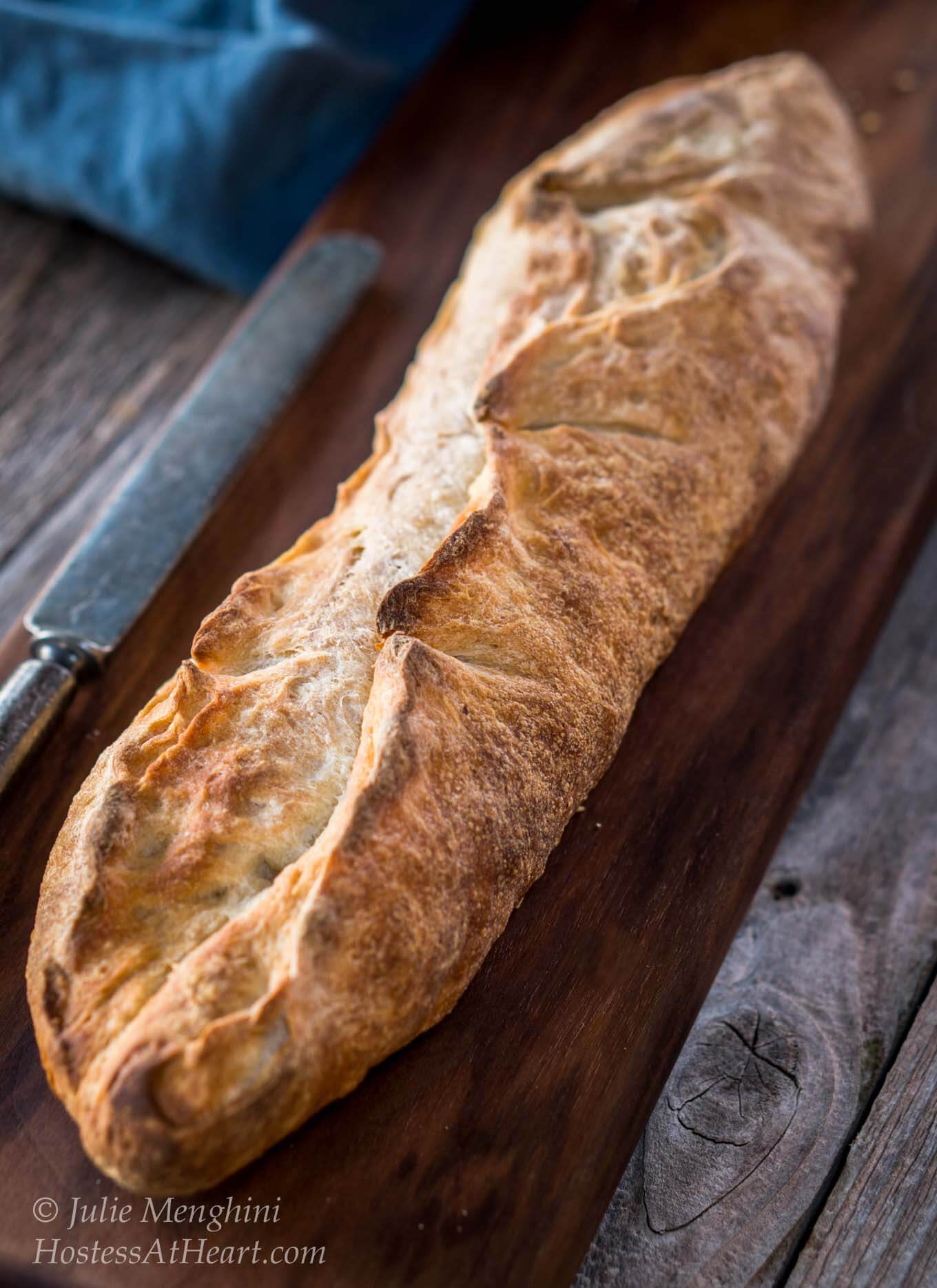 A baguette of bread sitting on a wooden cutting board. A knife and blue napkin sit next to the board.