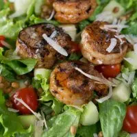 Close-up view of a white plate filled with a green salad, cucumbers, and tomatoes then topped with grilled shrimp and garnished with cheese. 