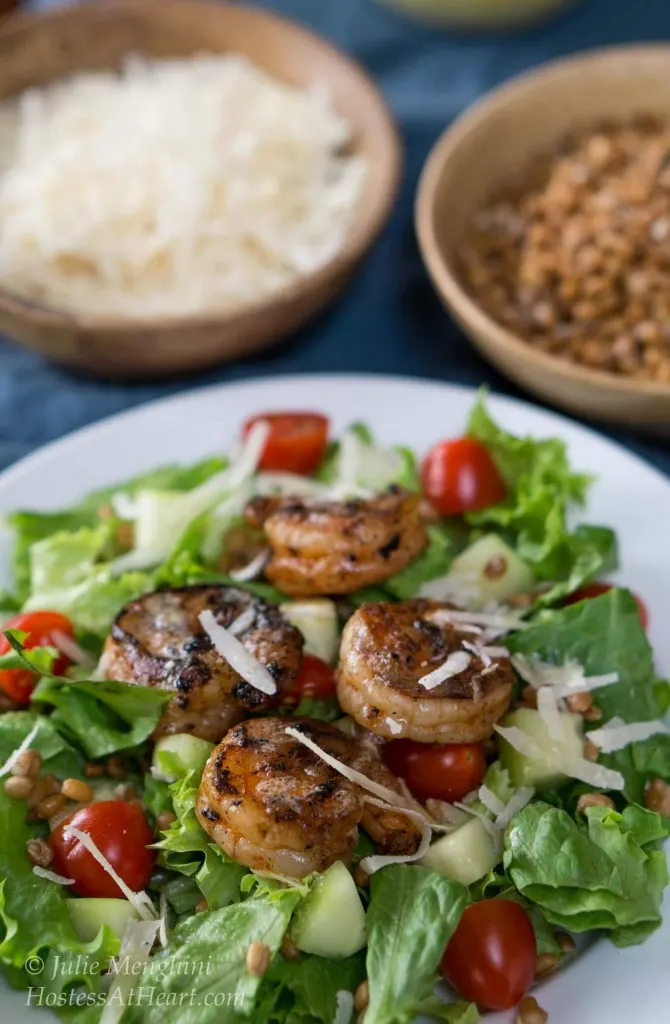 A white plate filled with a green salad, cucumbers, and tomatoes then topped with grilled shrimp and garnished with cheese. Wooden bowls of grated cheese and wheat berries sit in the background.
