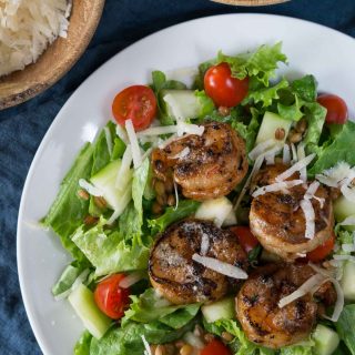 A white plate filled with greens, tomatoes, cucumbers, and grilled shrimp then garnished with shredded cheese.