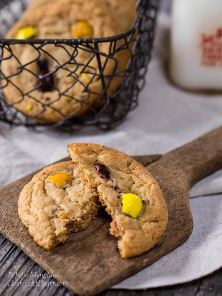 A broken M&M cookie sits on an antique butter paddle. A basket of cookies and a milk bottle sits in the background.