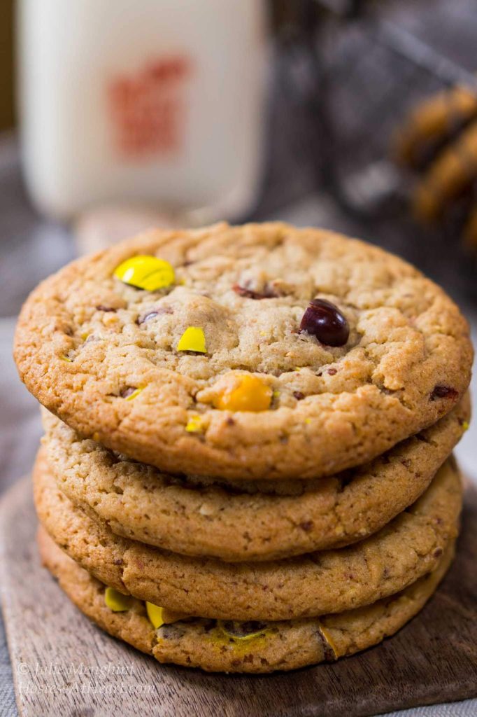 A stack of M&M cookies sitting on an antique butter paddle. A basket of cookies and a bottle of milk sit in the background.