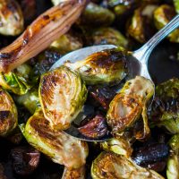 Top-down view of roasted brussels sprouts sitting on a serving spoon hovering over roasted Brussels sprouts with shallots and mushrooms.