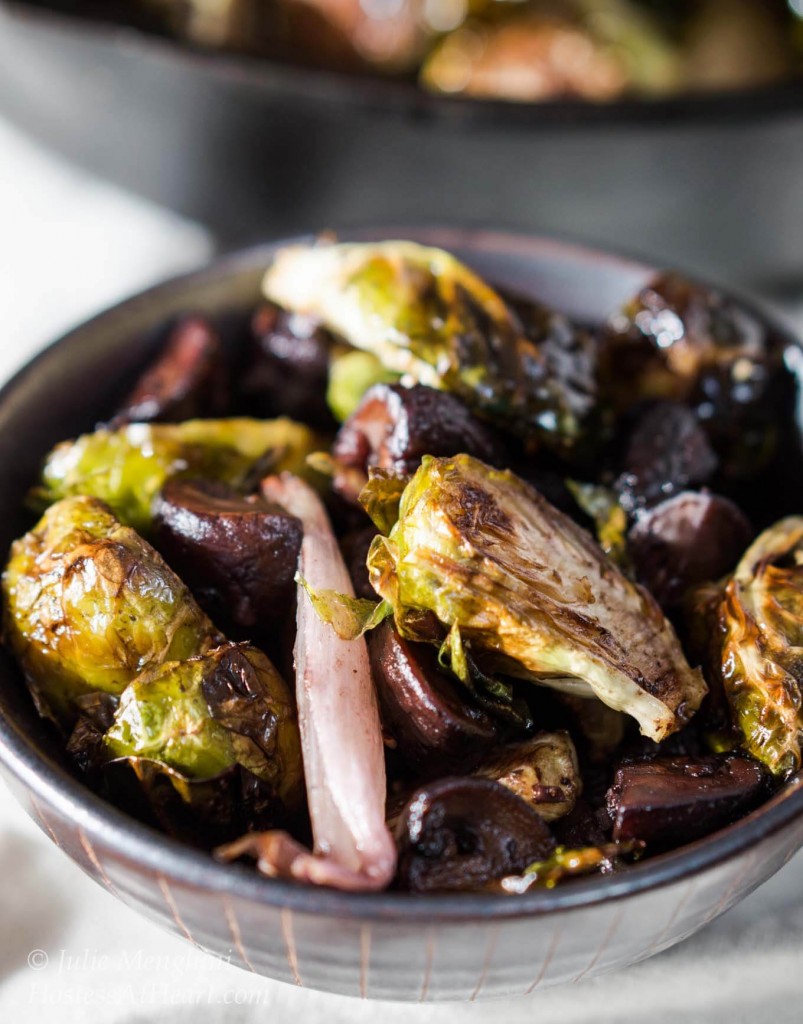 Top-down view of a brown bowl of roasted Brussels sprouts with shallots and mushrooms. The pan of Brussels Sprouts sit in the background.