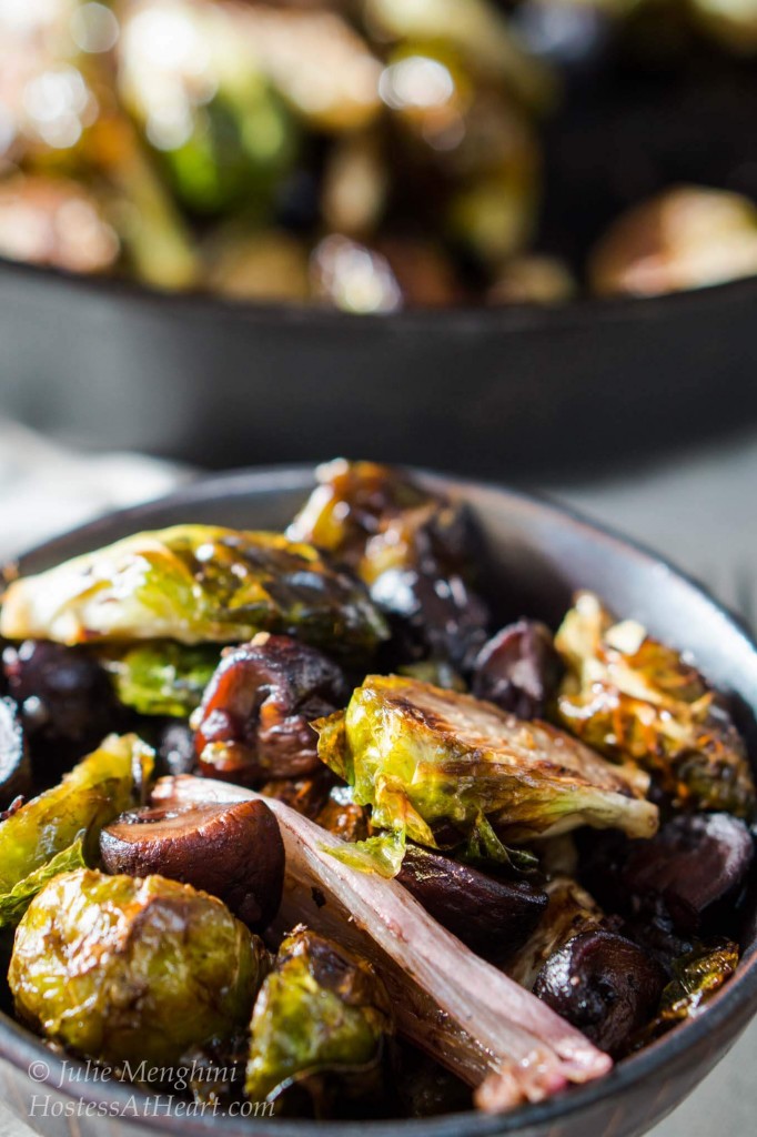 Top-down view of a brown bowl of roasted Brussels sprouts with shallots and mushrooms. The pan of Brussels Sprouts sits in the background.