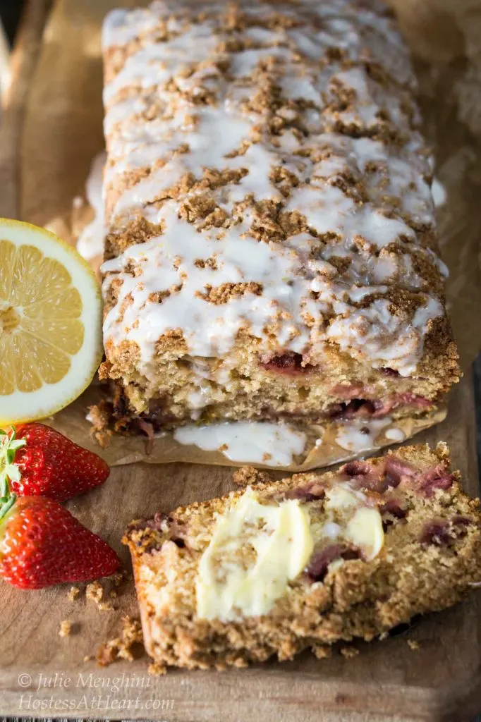 Top view of a loaf of Strawberry Rhubarb Bread with a streusel top that\'s drizzled with a glaze. The front piece has been cut from the loaf and is laying flat and covered in butter. Two strawberries and a slice of lemon sit to the side.