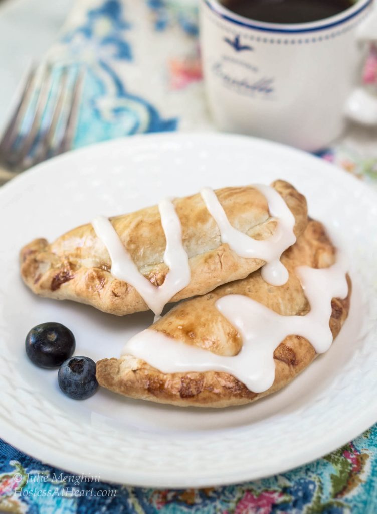 Two glazed pie crust hand pies filled with blueberries and pineapple sit on a white plate with two fresh berries. A fork and a cup of coffee sits in the back.
