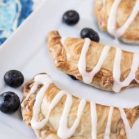 Pie crust crescents filled with Blueberry Pineapple filling on a white plate. Fresh Blueberries are scattered about. The crescents sit on a white plate over a colorful napkin.