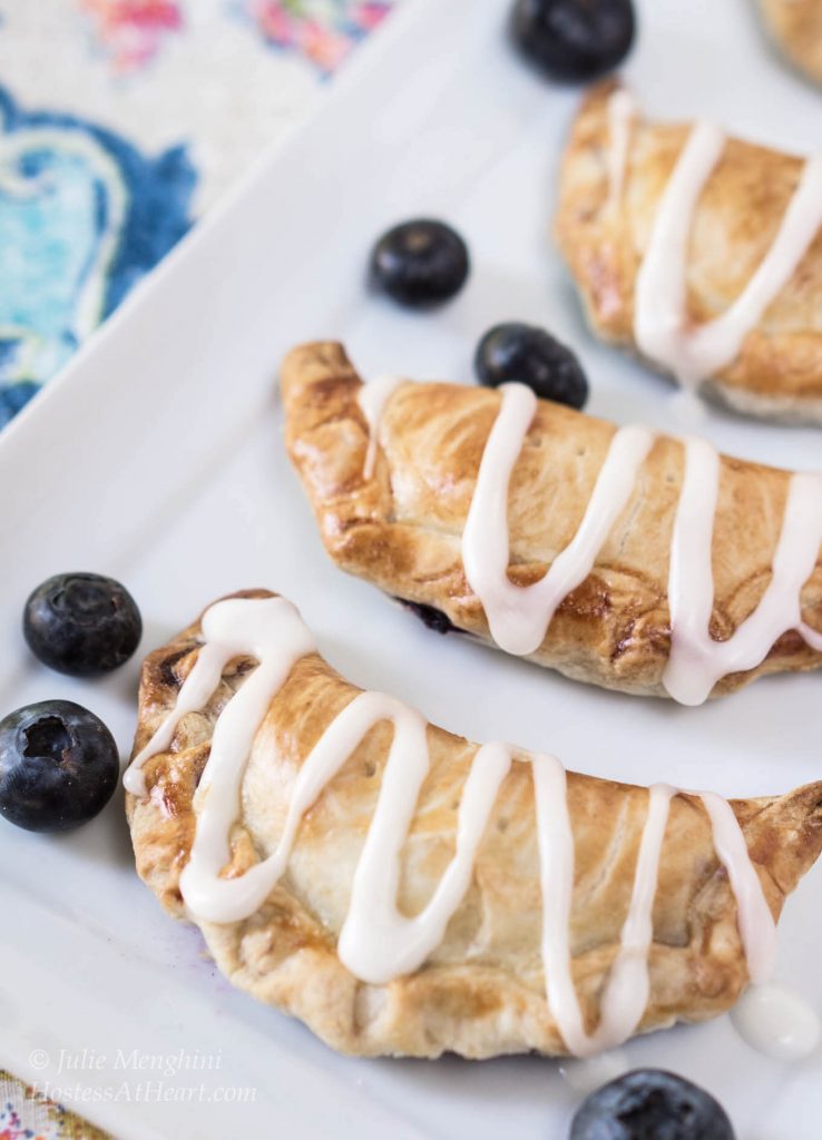Pie crust crescents filled with Blueberry Pineapple filling on a white plate. Fresh Blueberries are scattered about. The crescents sit on a white plate over a colorful napkin.