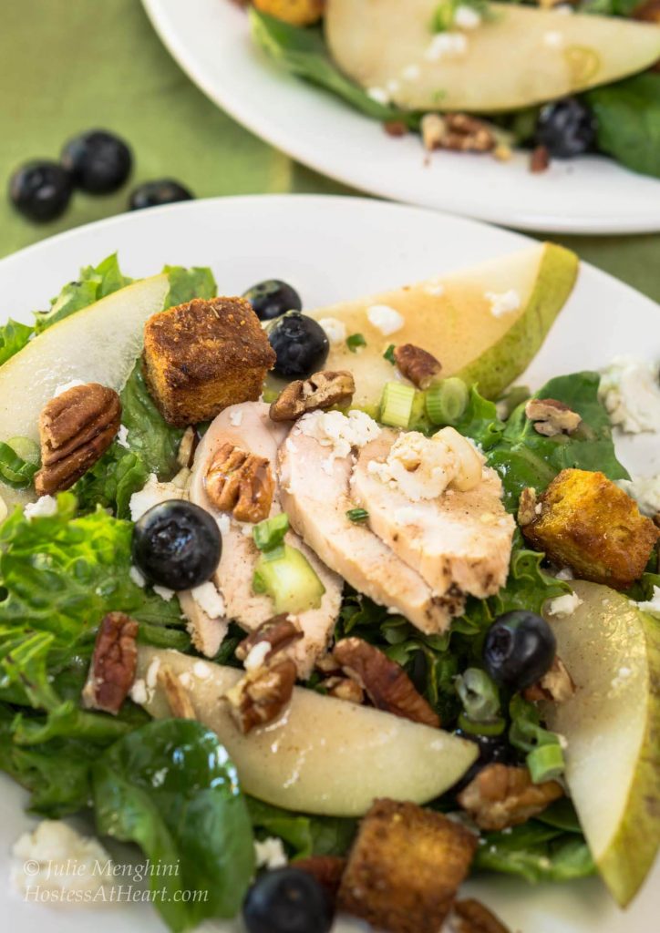 Green lettuce salad topped with sliced chicken, fresh pears, candied pecans, green onions, blueberries, and blue cheese crumbles. A second plate sits in the background.