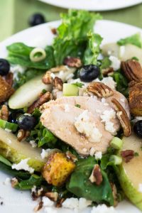 A plate of lettuce topped with sliced chicken blueberries, fresh slices of pears, candied pecans, crumbled blue cheese, and sliced green onions.