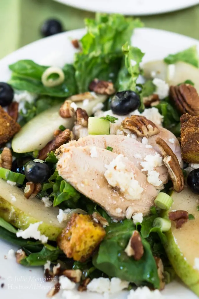 A close up of a plate filled with salad, fresh pears and blueberries, pecans, goat cheese, and sliced chicken.