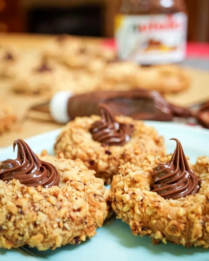 A close up a hazelnut thumbprint cookie filled with a swirl of Nutella. Cookies, a jar of Nutella, and a piping bag are in the background.