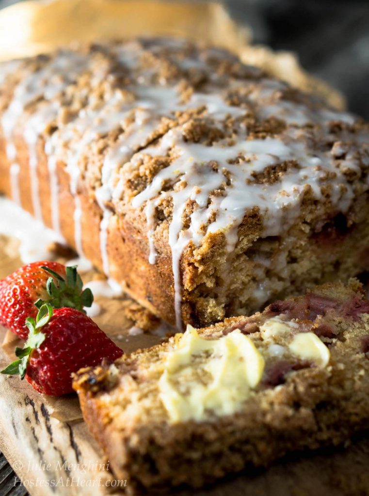 Side angle view of a loaf of Strawberry Rhubarb Bread topped with streusel and a drizzle. The front of the loaf is sliced with the slice laying on its side and spread with butter. Two strawberries sit next to the loaf.