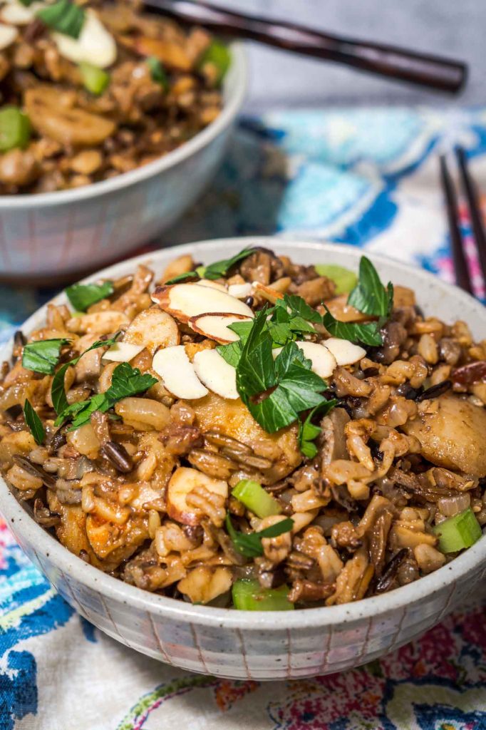 A front view of a gray bowl filled with 5 Spice Rice garnished with sliced almonds, sliced green onions, and chopped parsley. A second bowl sits in the background and a pair of chopsticks sit to the side over a colorful napkin.