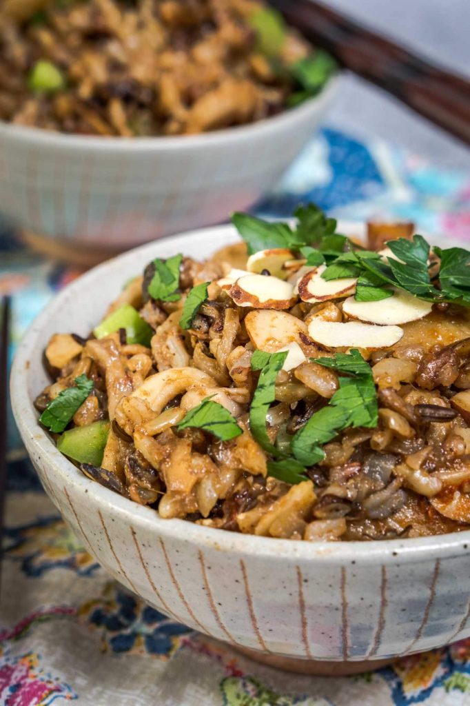 A front view of a gray bowl filled with 5 Spice Rice garnished with sliced almonds, sliced green onions, and chopped parsley. A second bowl sits in the background over a colorful napkin.