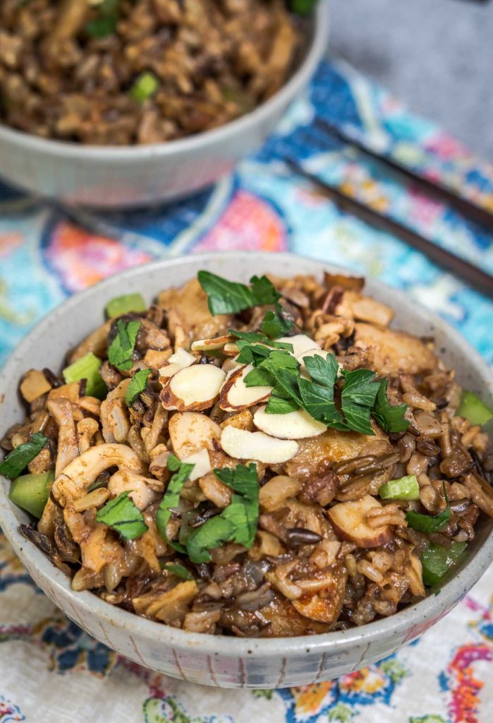 A front view of a gray bowl filled with 5 Spice Rice garnished with sliced almonds, sliced green onions, and chopped parsley. A second bowl sits in the background and a pair of chopsticks sit to the side over a colorful napkin.