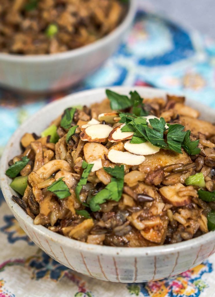 A front view of a gray bowl filled with 5 Spice Rice garnished with sliced almonds, sliced green onions, and chopped parsley. A second bowl sits in the background and a pair of chopsticks sit to the side over a colorful napkin.