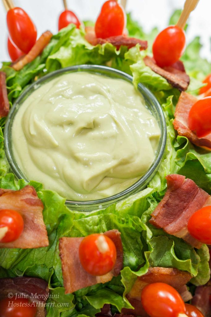 Front down photo of a head of lettuce that\'s been hollowed out and filled with Avocado sauce. Skewers filled with bacon and tomatoes stick out from the lettuce.