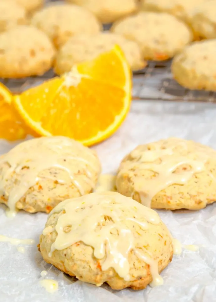 Front view of Citrus Carrot Cookies topped with glaze sit on parchment paper. Slices of oranges and a cooling rack of the cookies sit in the background.
