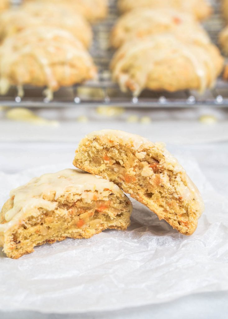 A Citrus Carrot Cookie cut in half and stacked together over a piece of parchment paper. A cooling rack of the glazed cookies sits in the background.