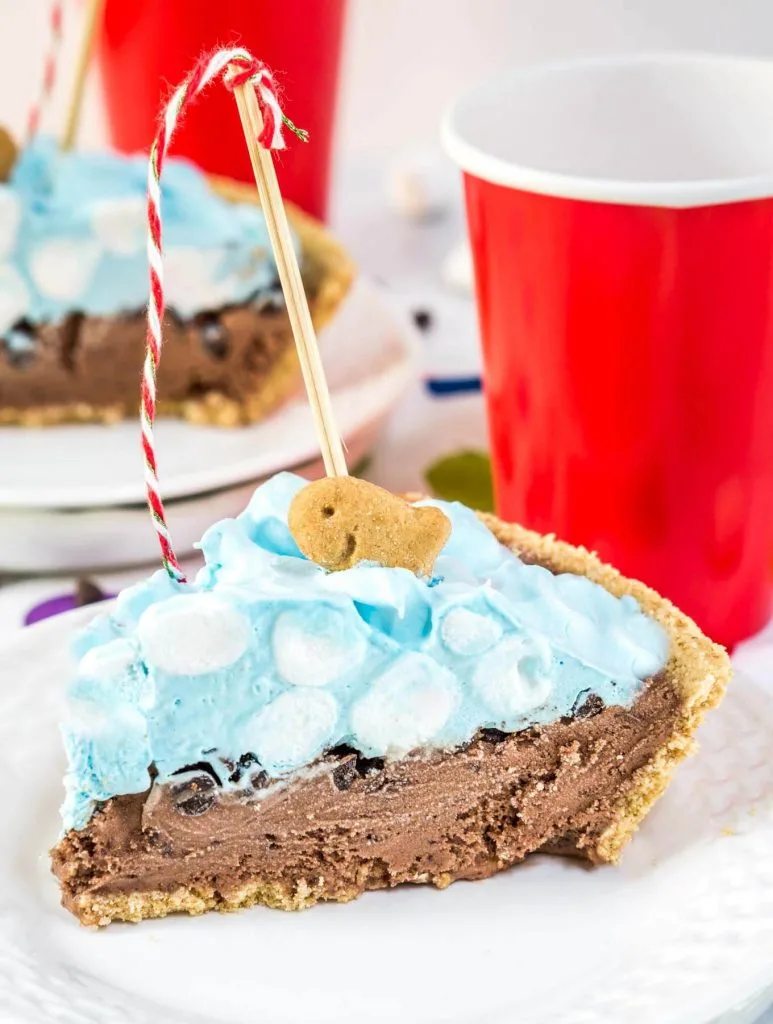 A slice of ice cream pie designed like a swimming pool sitting on a white plate over a tablecloth with colorful circles on it. A crust is filled with chocolate filling topped with blue-hued cool whip and mini marshmallows. A fishing pole with a red striped string is attached to a graham cracker fish. A second slice sits in the background. Red cups sit next to each slice of pie.
