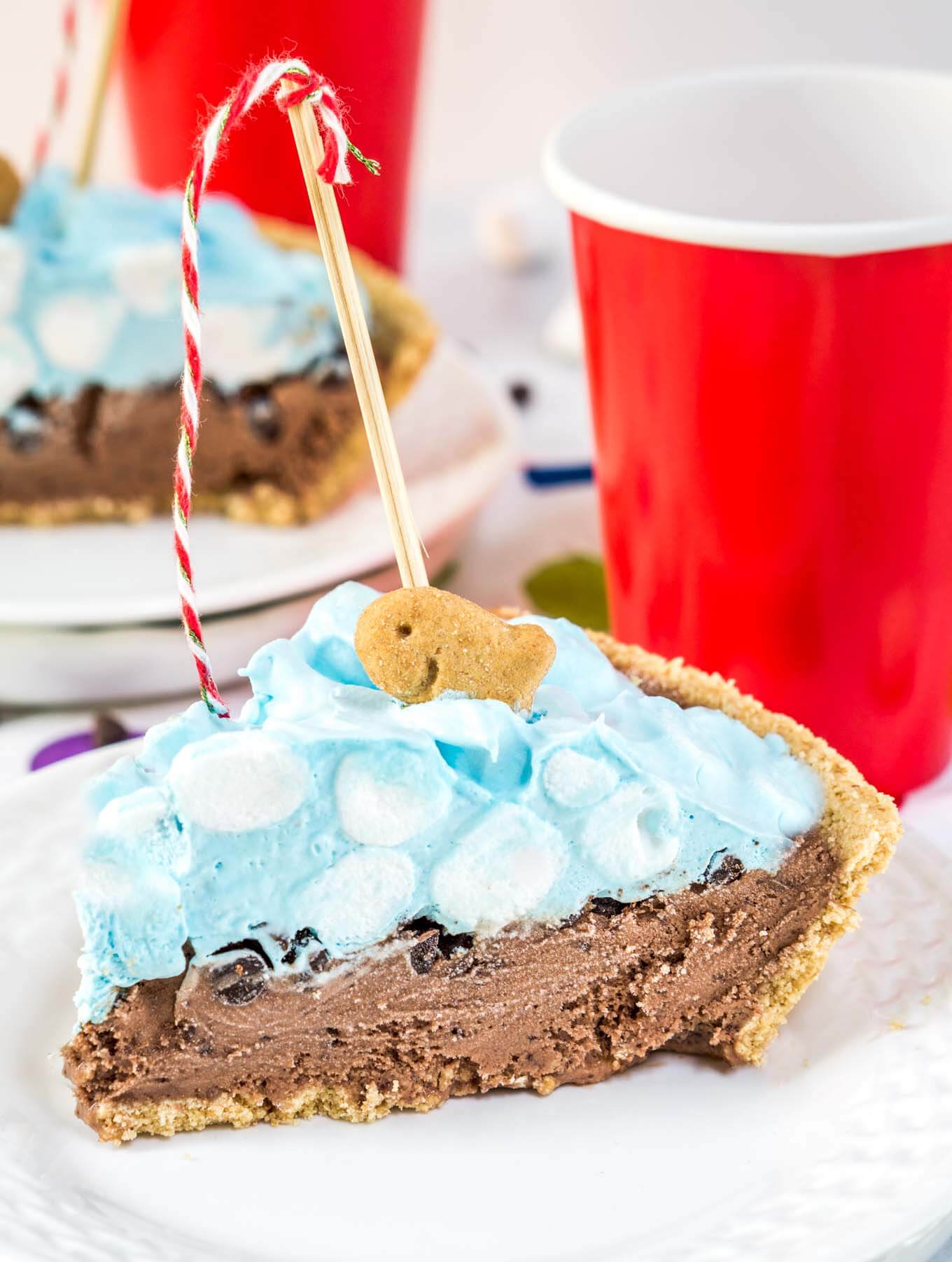 A slice of ice cream pie designed like a swimming pool sitting on a white plate over a tablecloth with colorful circles on it. A crust is filled with chocolate filling topped with blue-hued cool whip and mini marshmallows. A fishing pole with a red striped string is attached to a graham cracker fish. A second slice sits in the background. Red cups sit next to each slice of pie.