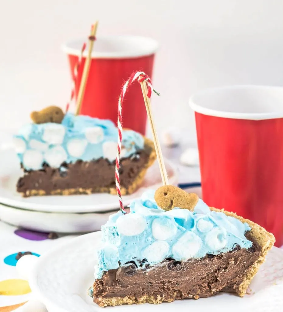 A slice of ice cream pie designed like a swimming pool sitting on a white plate over a tablecloth with colorful circles on it. A crust is filled with chocolate filling topped with blue-hued cool whip and mini marshmallows. A fishing pole with a red striped string is attached to a graham cracker fish. A second slice sits in the background. Red cups sit next to each slice of pie.