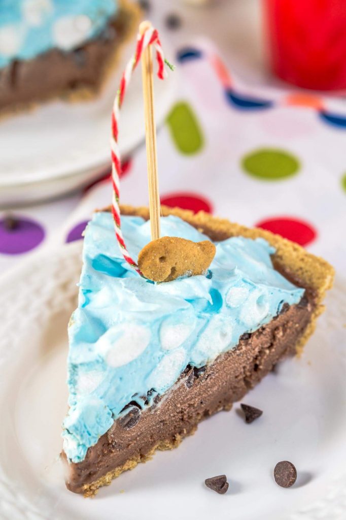 A slice of ice cream pie designed like a pool sitting on a white plate over a tablecloth with colorful circles on it. A crust is filled with chocolate filling topped with blue-hued cool whip and mini marshmallows. A fishing pole with a red striped string is attached to a graham cracker fish. A second slice sits in the background.