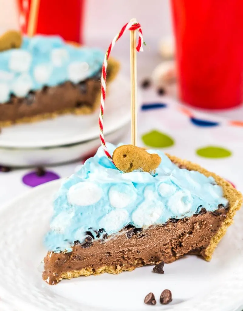 A slice of ice cream pie topped with blue-colored ice cream and marshmallows indicating it\'s water. A goldfish cookie with on the top with a crafted fishing pole. A second piece sits in the backgroud.