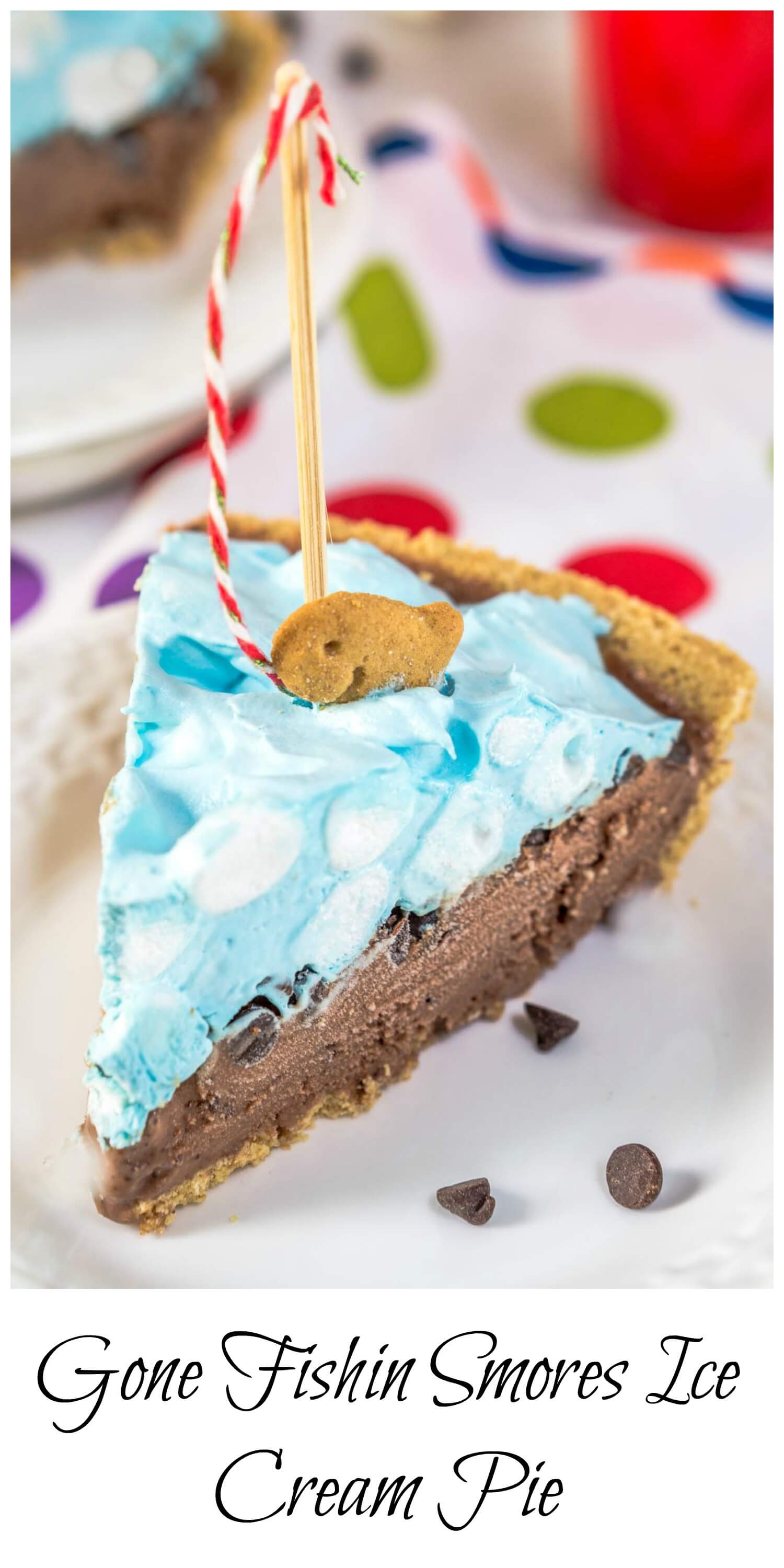 A slice of ice cream pie designed like a swimming pool sitting on a white plate over a tablecloth with colorful circles on it. A crust is filled with chocolate filling topped with blue-hued cool whip and mini marshmallows. A fishing pole with a red striped string is attached to a graham cracker fish.