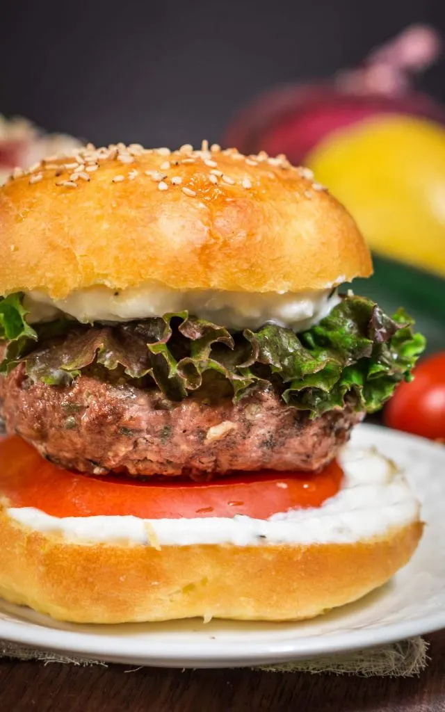 Lamb Burger between a sesame seed bun with a layer of Lemon Herb Sauce, red lettuce, and a slice of tomato sitting on a white plate.