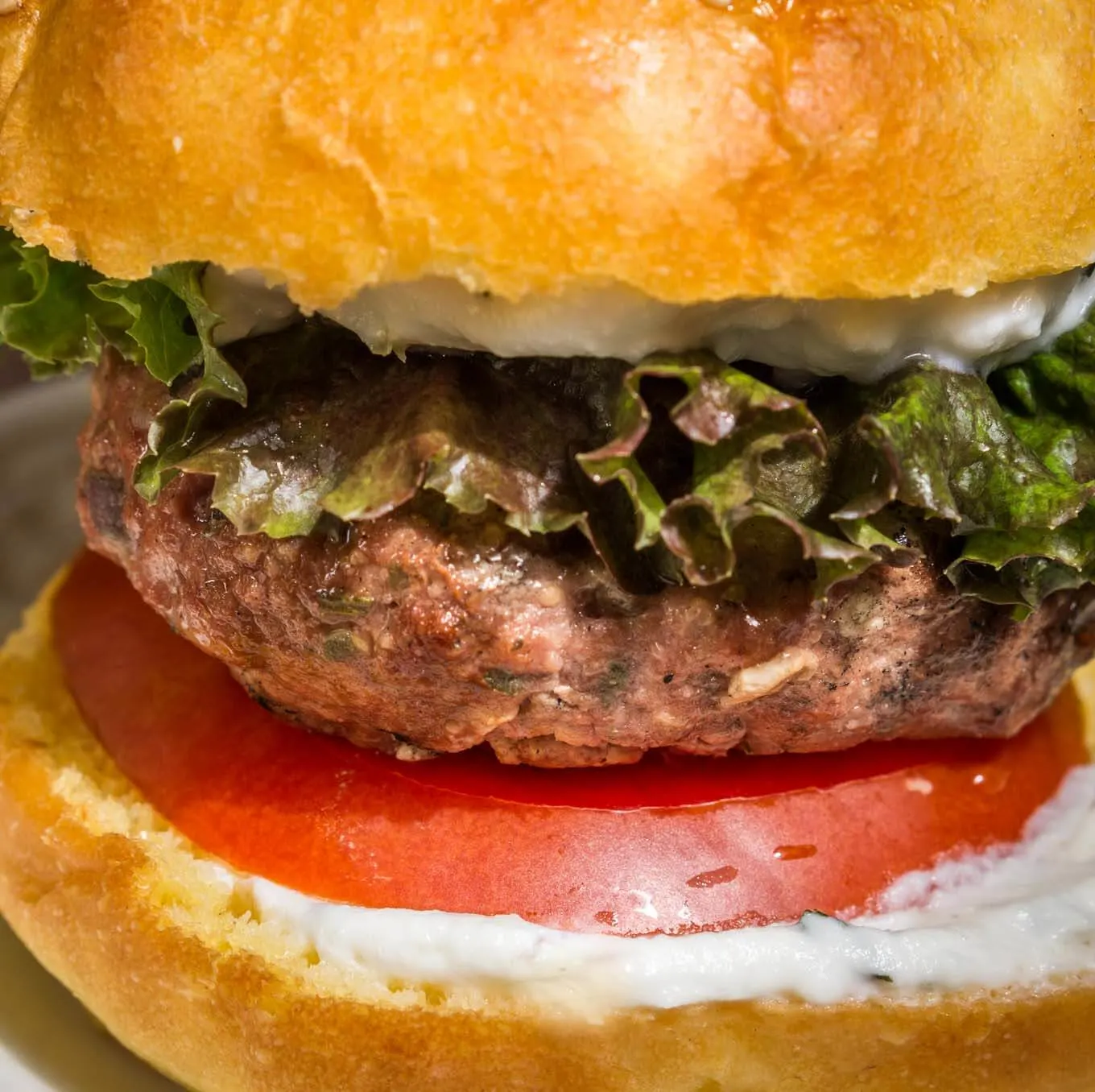 Sideview of a Greek Lamb Burger topped with a bun that\'s been layered with lemon herb sauce and lettuce.