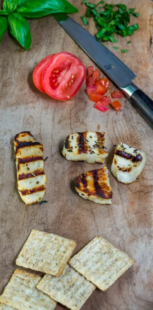 Grilled Halloumi cheese and diced tomatoes and knife sit on a wooden cutting board.